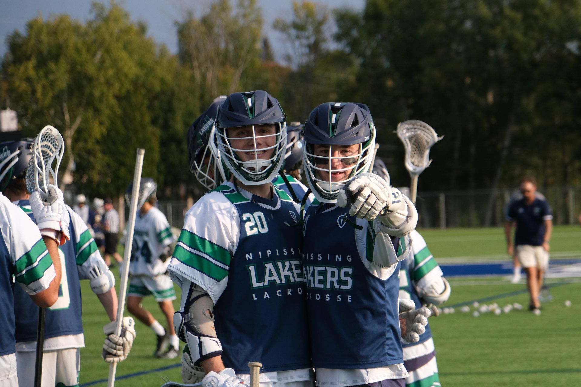 Two lacrosse players in navy and green jerseys smiling on a field.