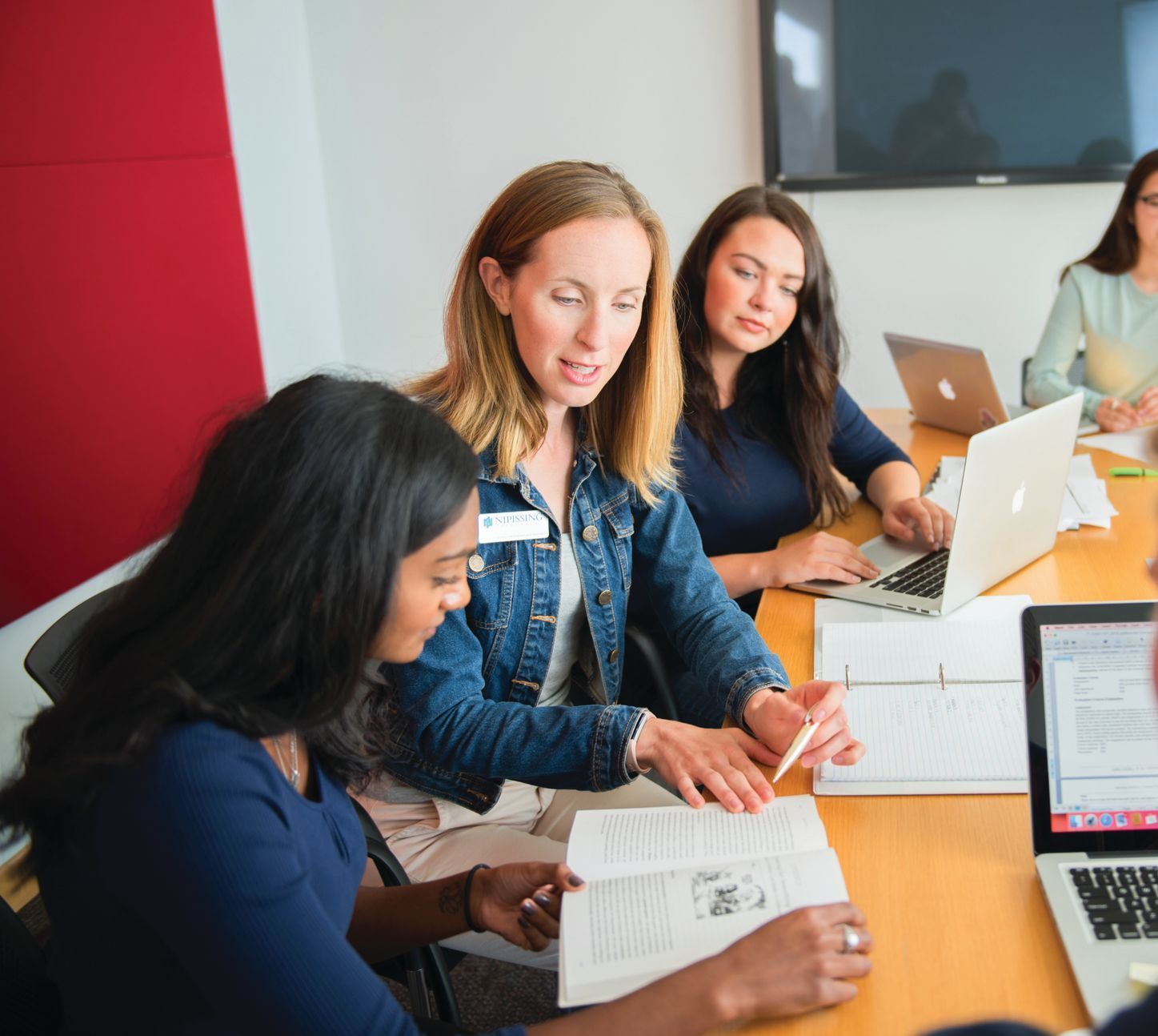 Four women in a meeting. A woman with a jean jacket points to a book while others look on.