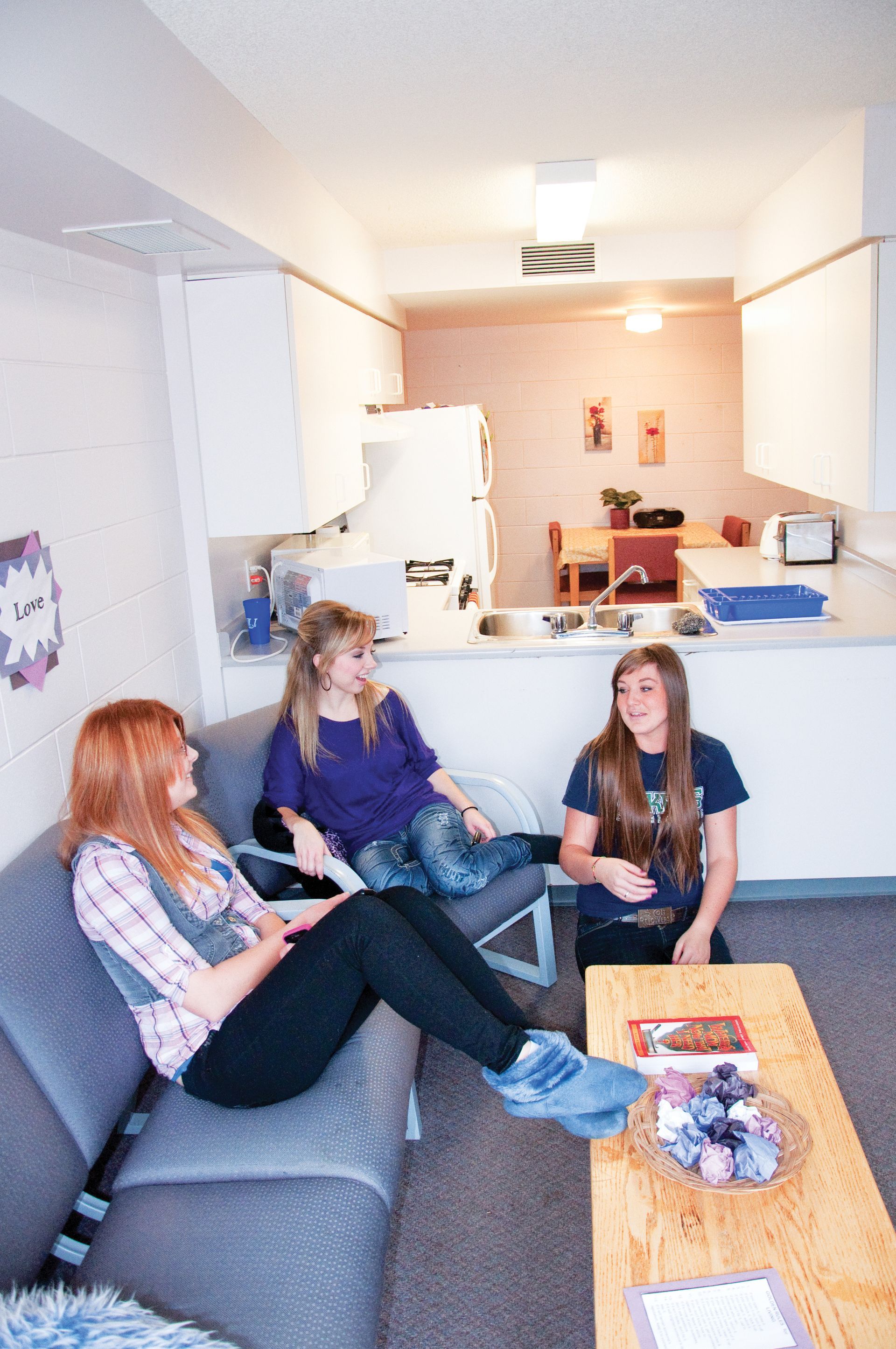 Three young women in a small apartment, conversing. One lounges on a couch, two seated nearby.