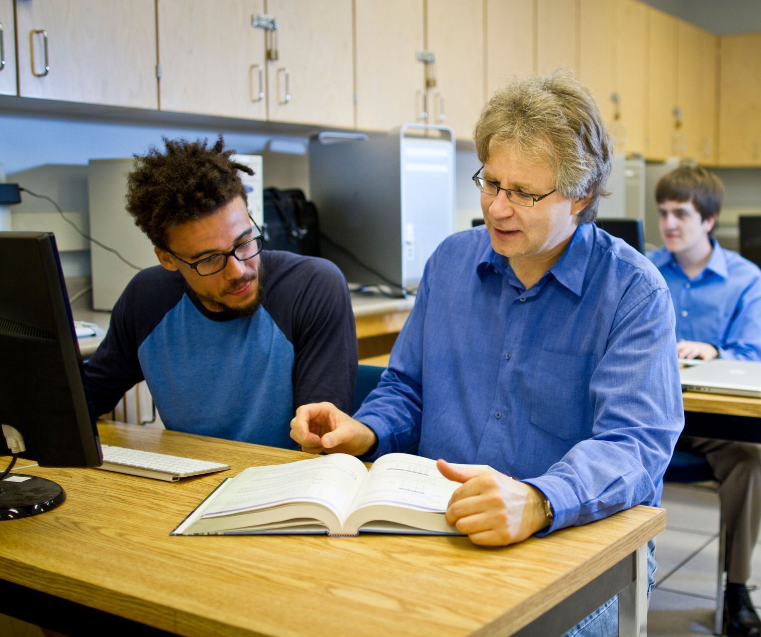 A professor helping a student at a computer, reading from a book, in a classroom.