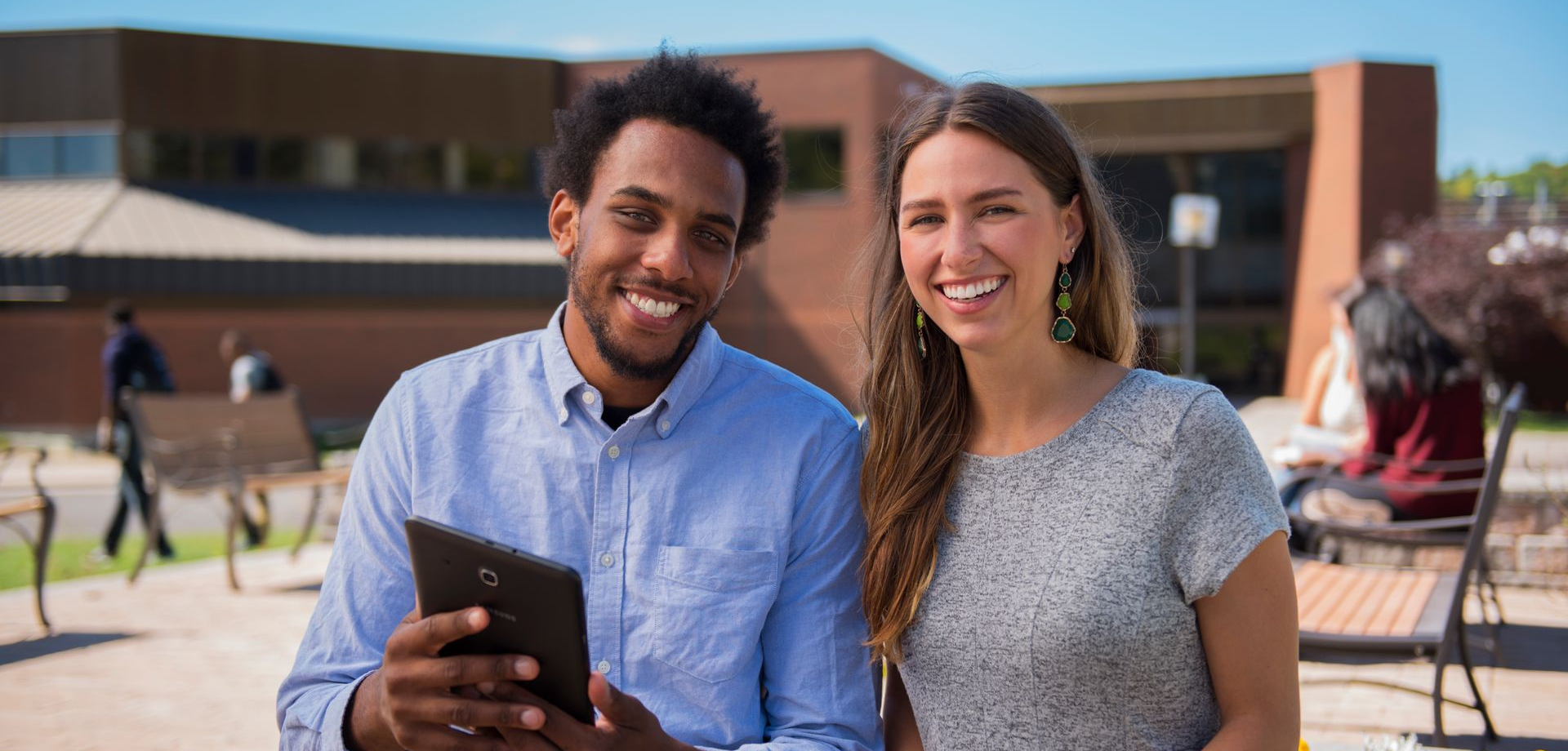 Two students smile while working on a tablet and laptop outside a brick building on a sunny day.