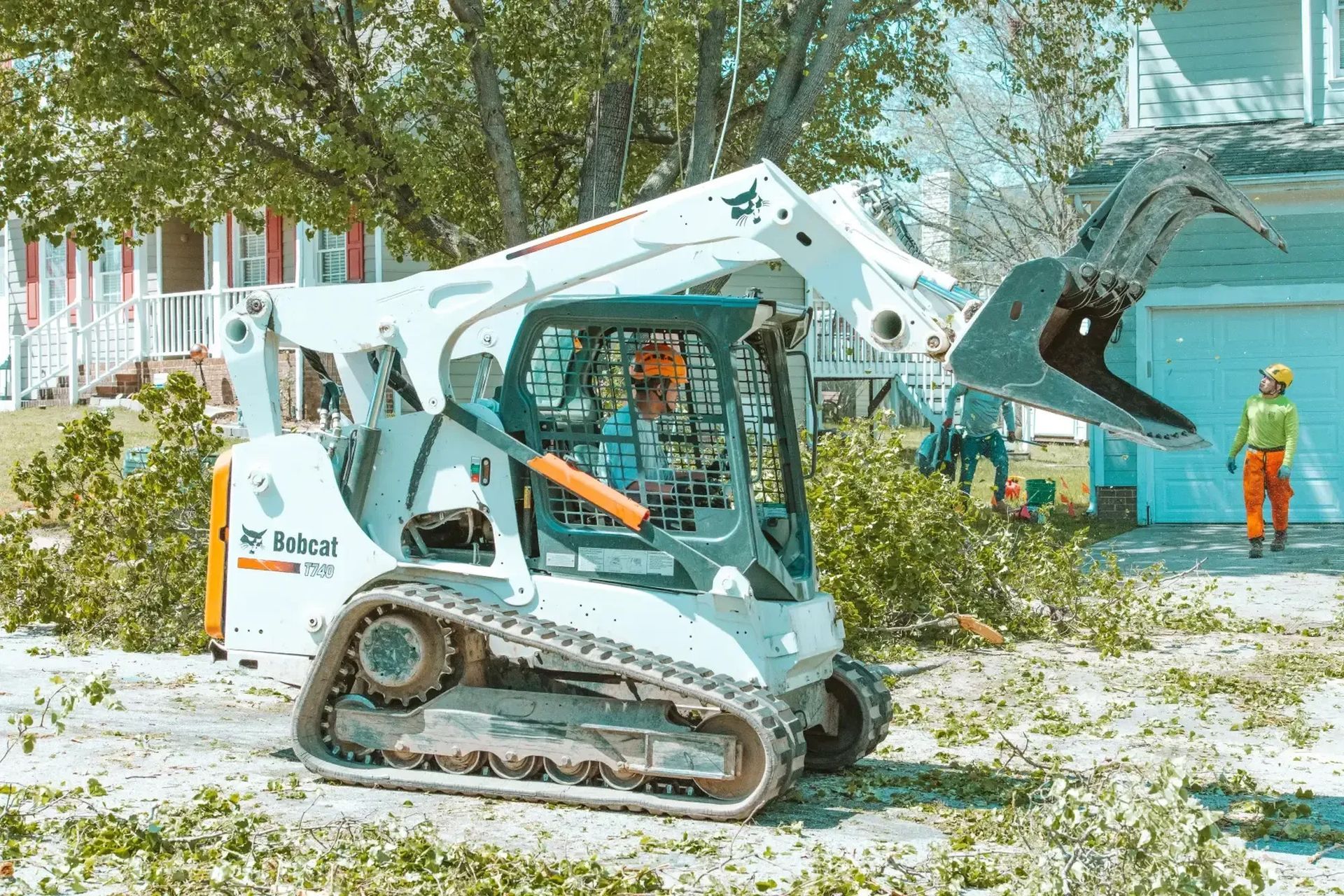 A bobcat is cutting trees in front of a house.