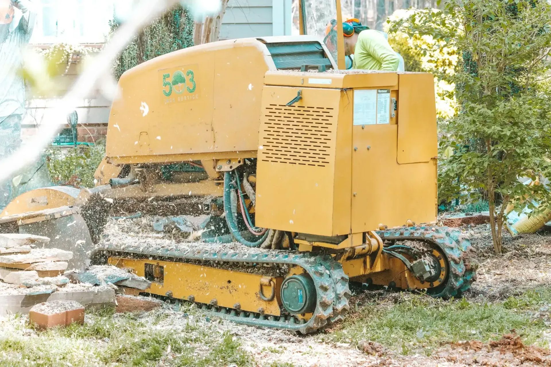 A yellow tractor is cutting a tree stump in a yard.
