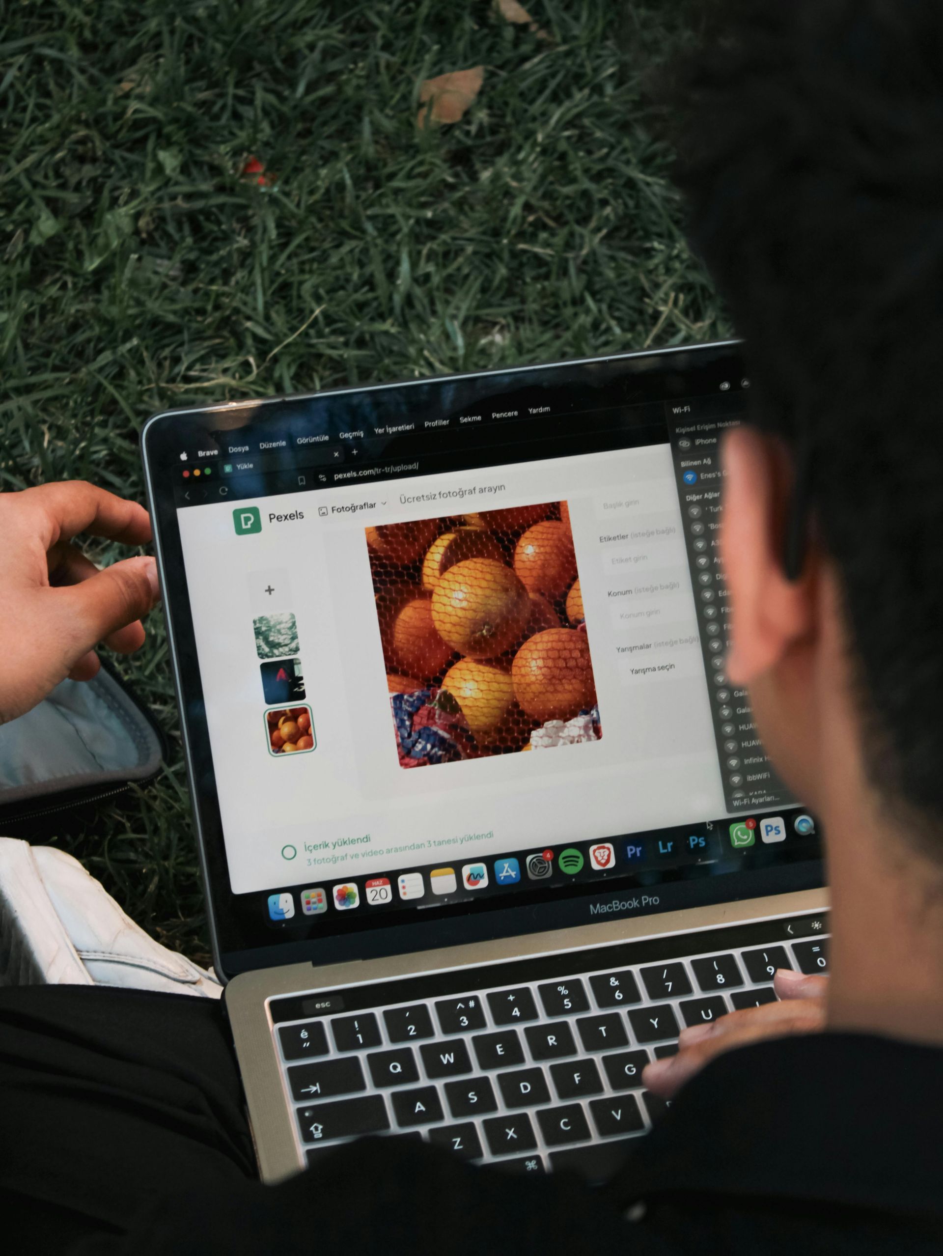 Person using a laptop on grass; an image of oranges is open on the screen, with editing tools visible.