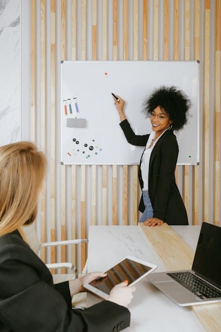 Woman presenting at a whiteboard, pointing with a marker. Another person uses a tablet at a table.