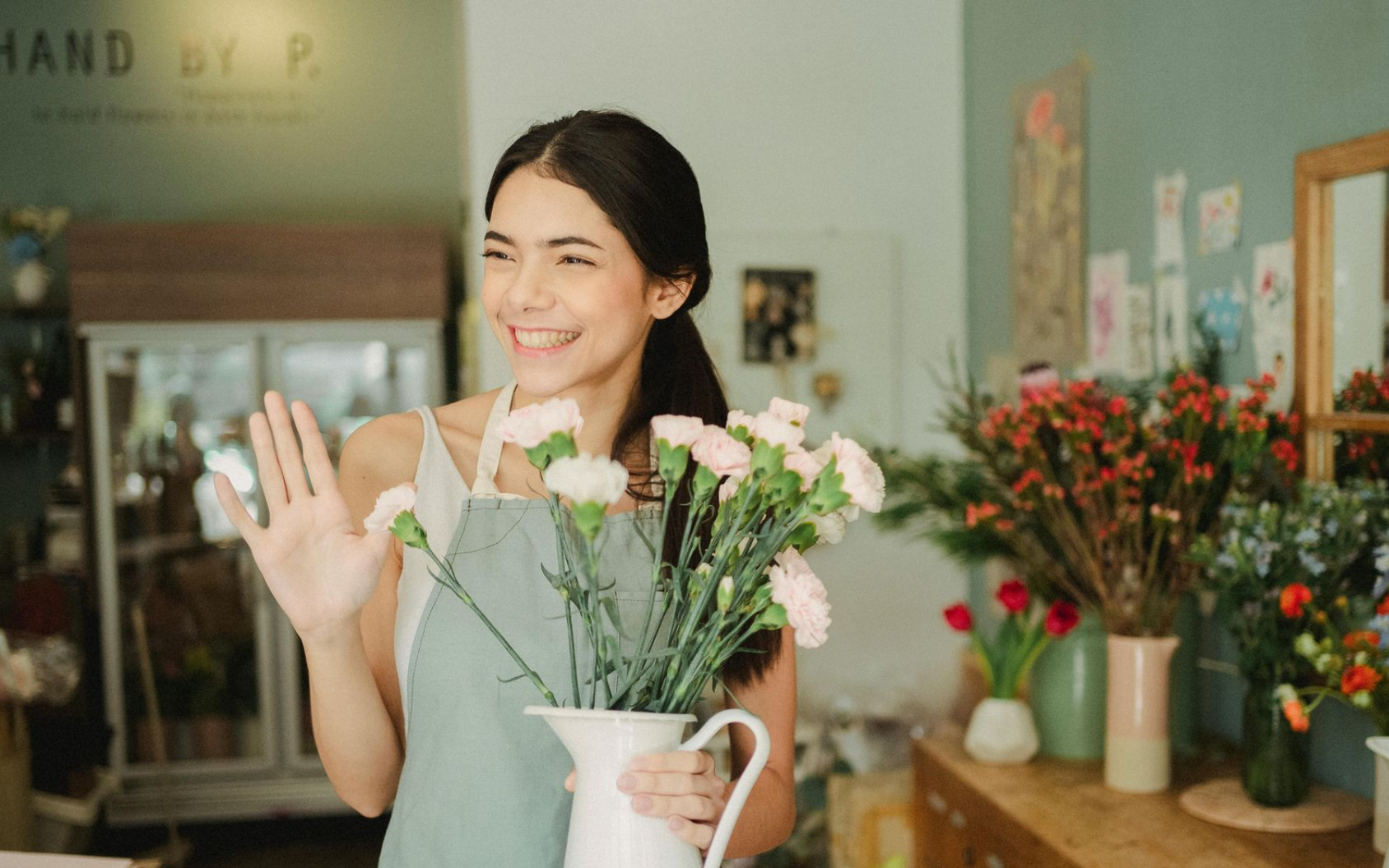 Small business owner in apron holding flowers, waving in a flower shop, smiling.