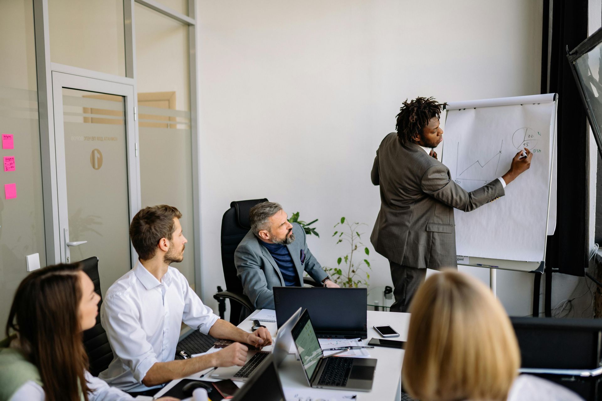 A person in a suit stands at a whiteboard, gesturing to a diagram, while colleagues watch during a meeting.