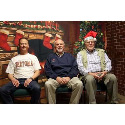 Three men are sitting next to each other in front of a christmas tree.