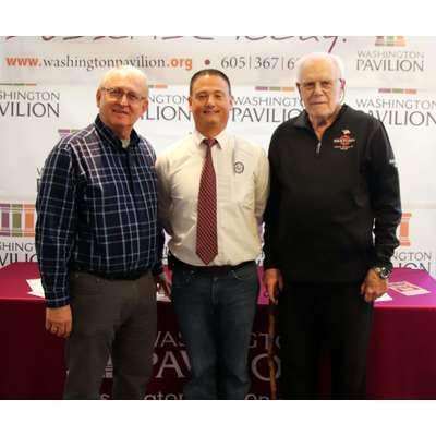 Three men are posing for a picture in front of a washington pavilion sign.