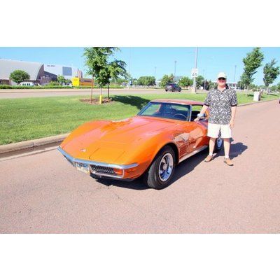 A man is standing next to an orange corvette in a parking lot.