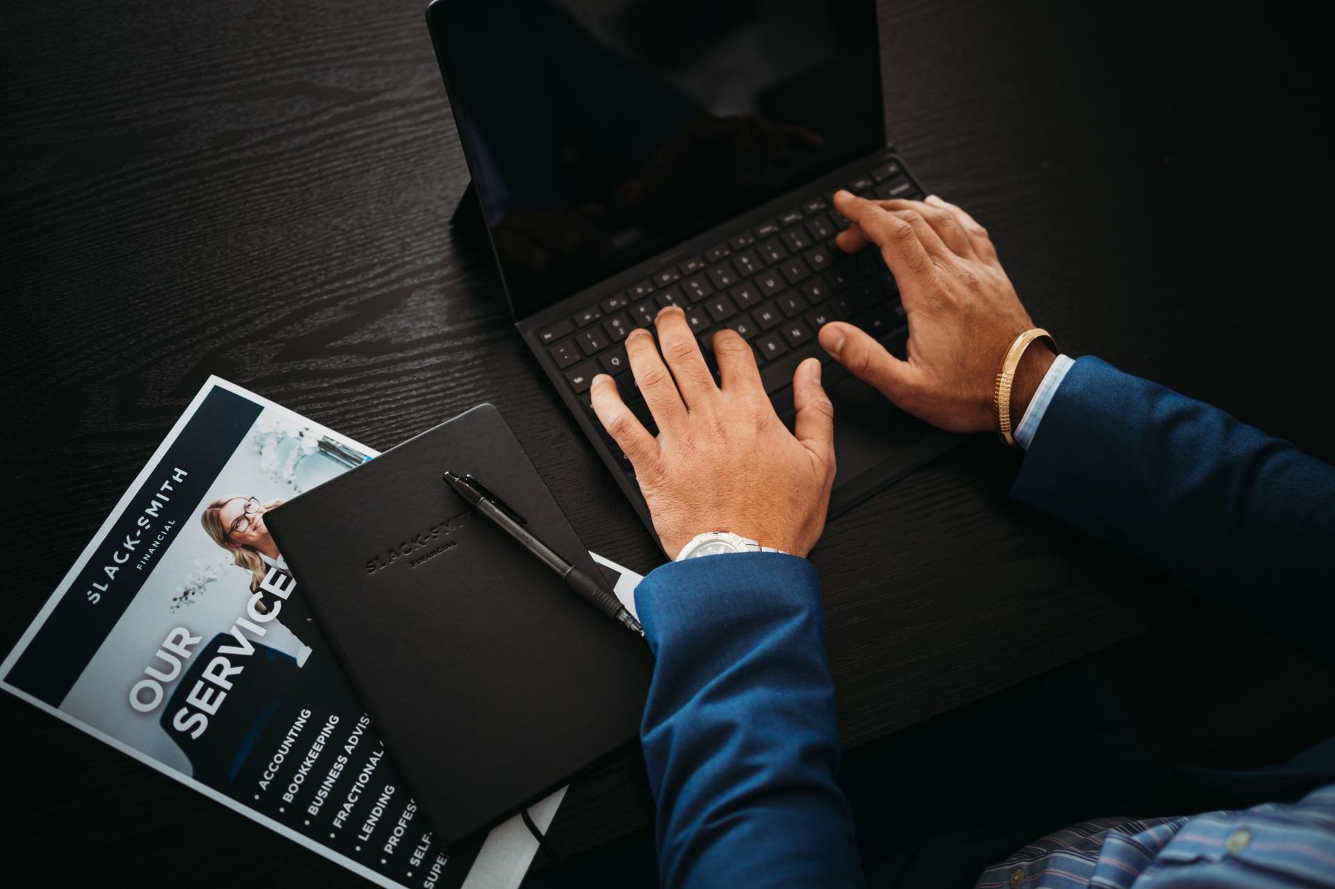 Person in blue suit typing on a laptop, with a notebook, pen, and brochure on a dark desk — Slack-Smith Partners in Tamworth, NSW 