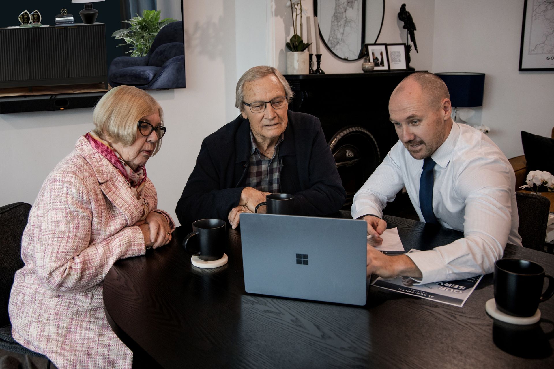 Older Couple and Advisor Looking at a Laptop, Discussing Documents at a Table. Indoors, Dark Wood Table, Black Mugs — Slack-Smith Partners In Moree, NSW