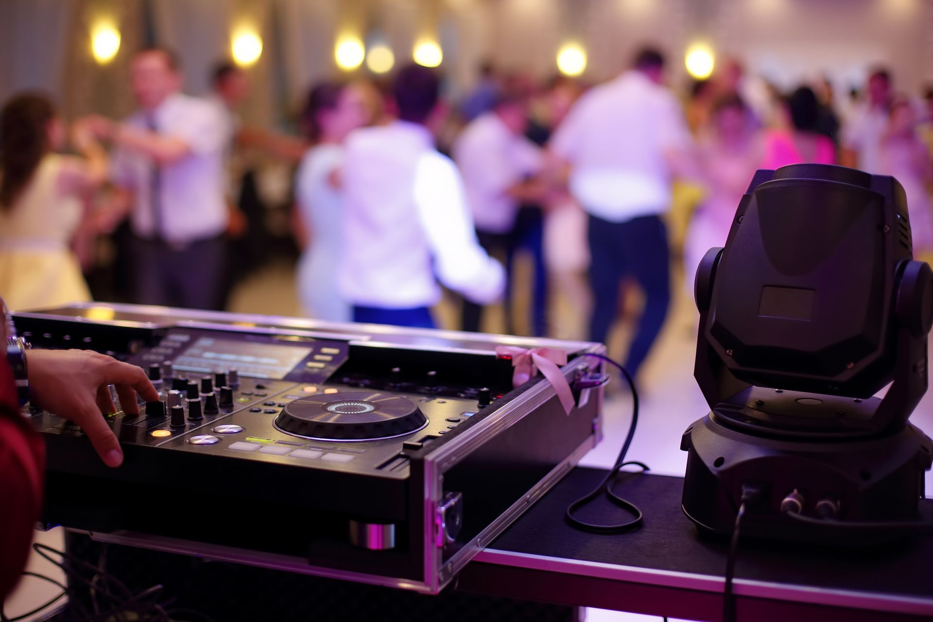 DJ mixing music at a party; people dancing in the background. Close-up of equipment and blurred figures.