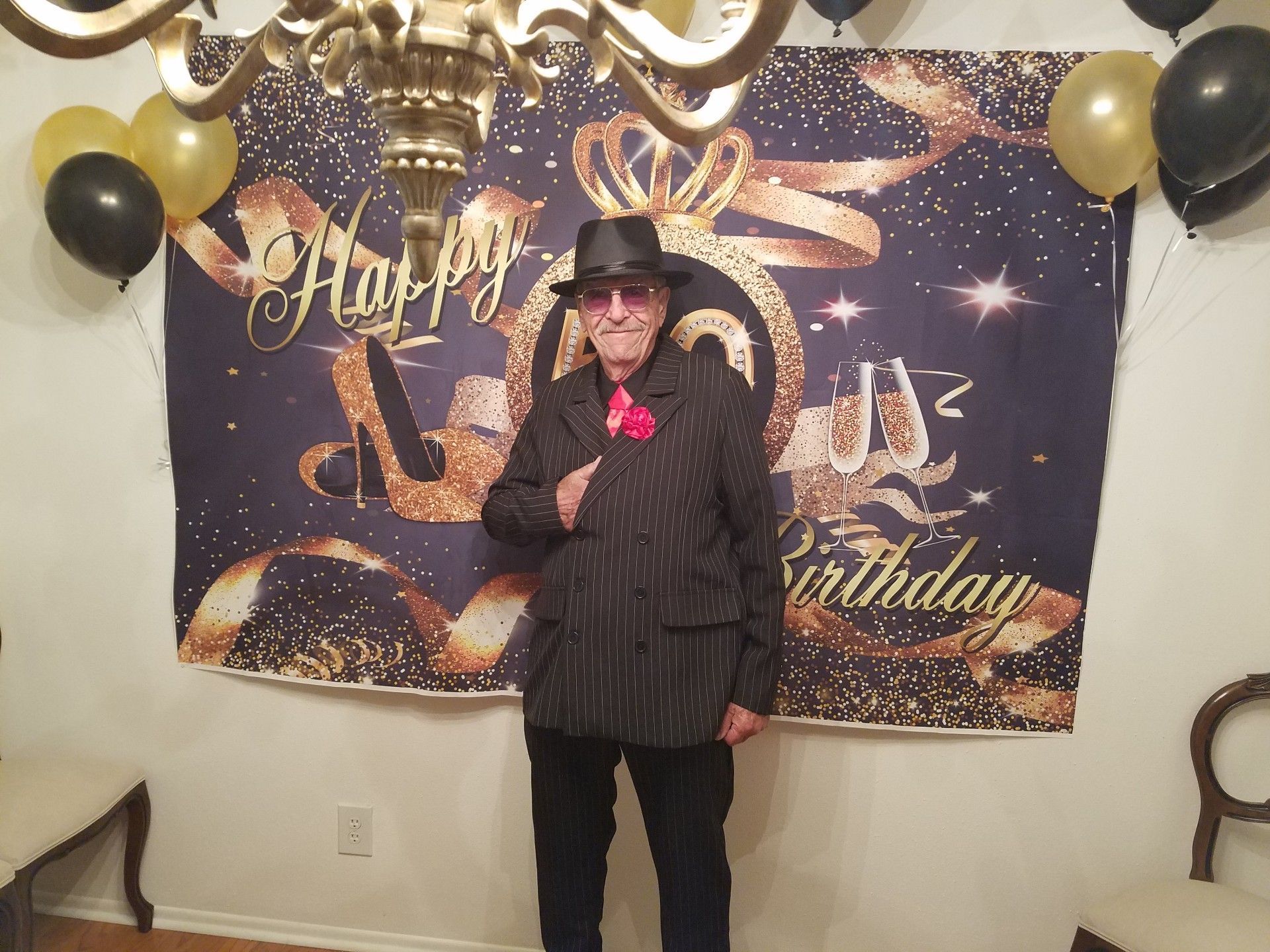 Man in suit and fedora poses in front of a birthday backdrop with balloons.