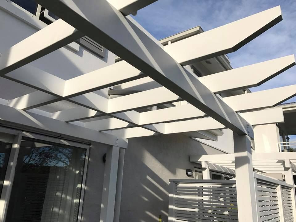 White pergola over an outdoor patio, casting shadows on the light gray building and fence.
