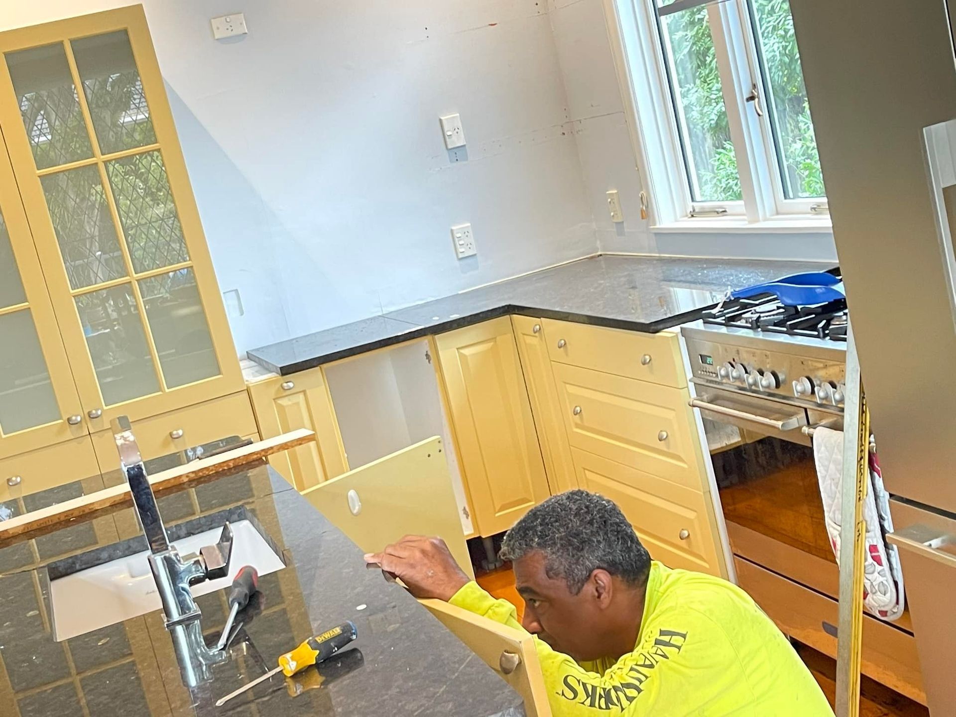 A person installing a cabinet in a kitchen, wearing a yellow shirt, with black countertops and a window.