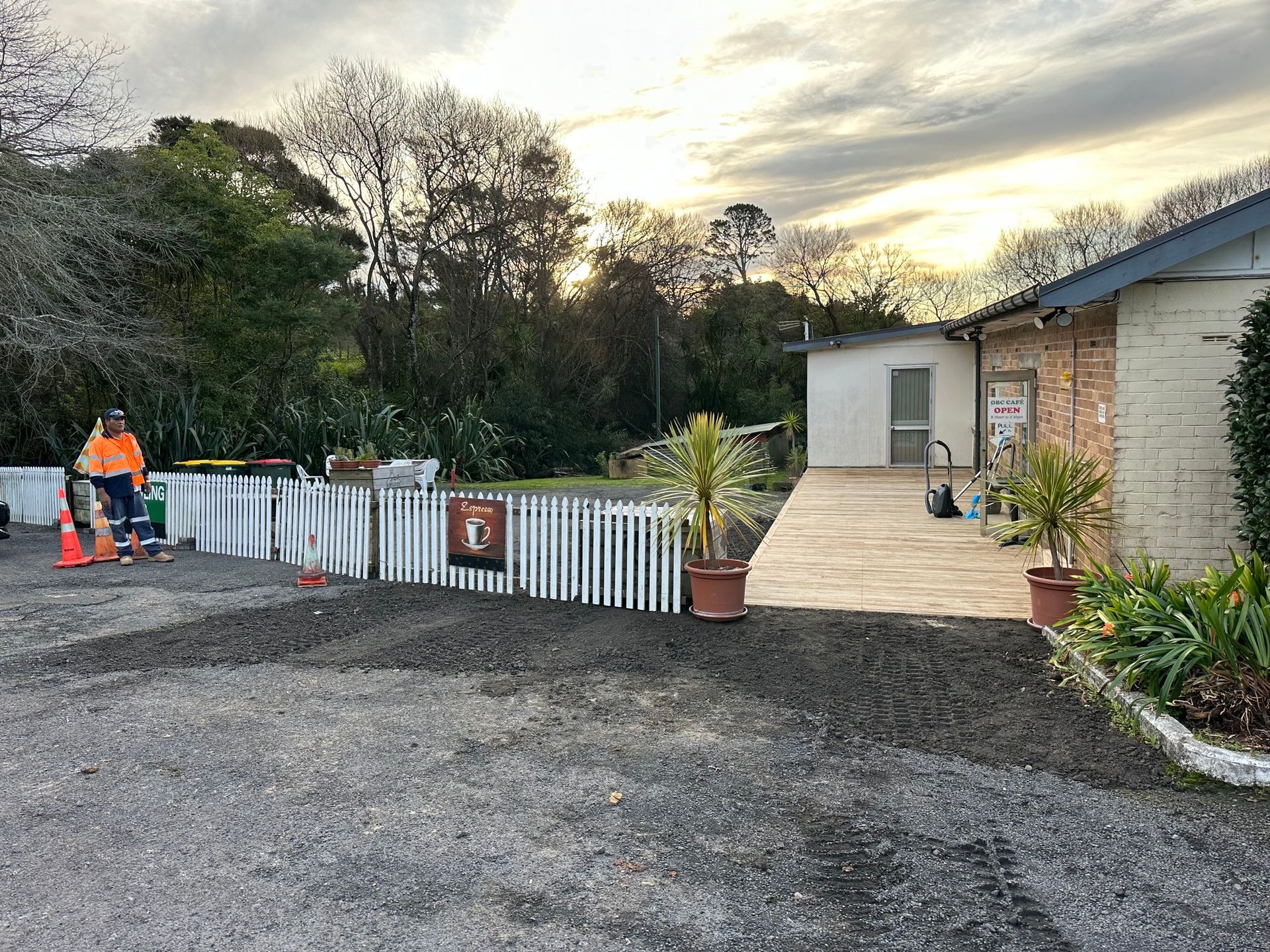 Gravel driveway leading to a building with a wooden walkway and potted plants. A person in orange safety gear operates a machine. Gravel driveway leading to a building with a wooden walkway and potted plants. A person in orange safety gear operates a machine.