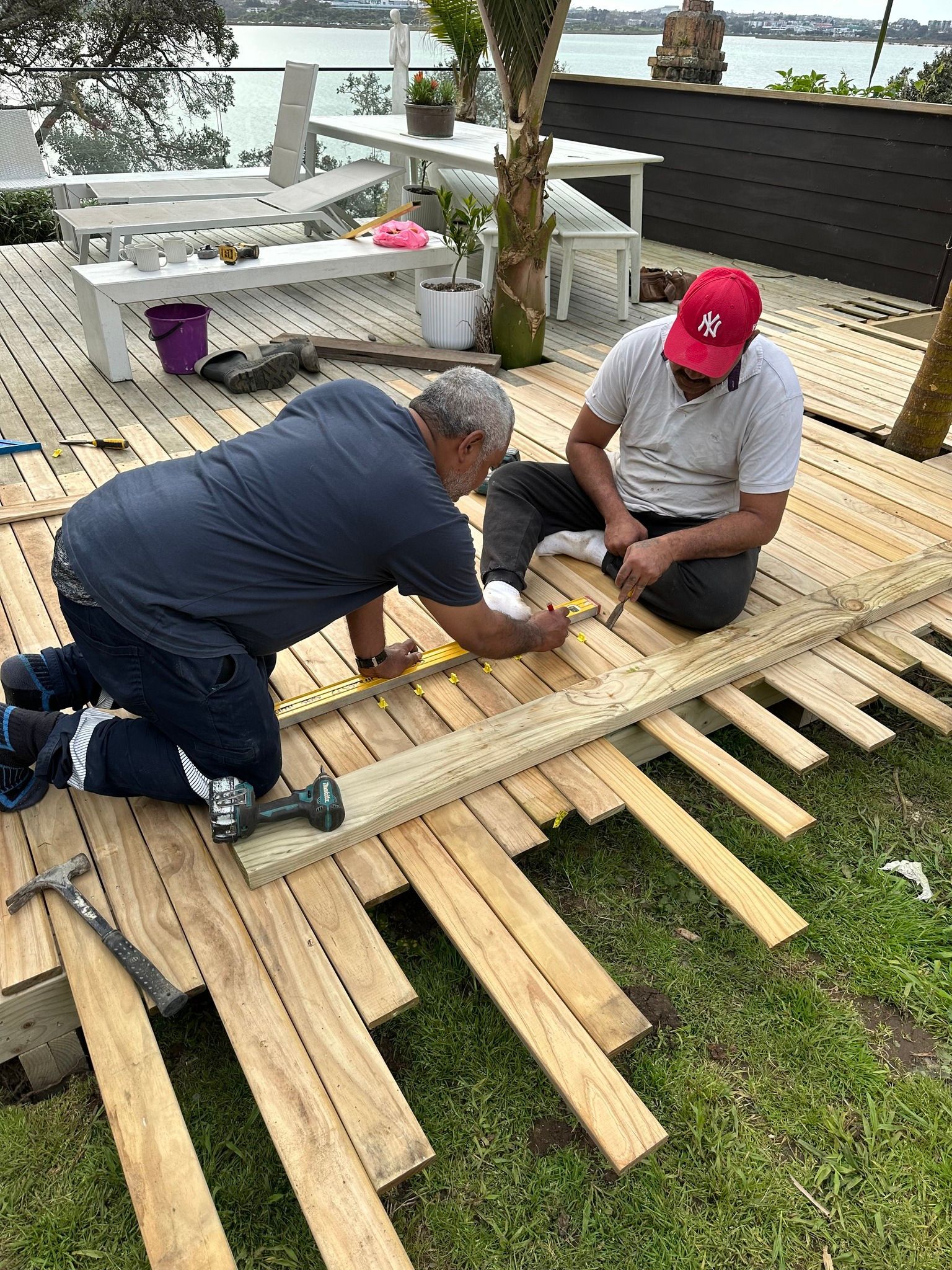 Two men building a wooden deck outdoors. One kneels and works with a tool. The other sits, also working. Two men building a wooden deck outdoors. One kneels and works with a tool. The other sits, also working.