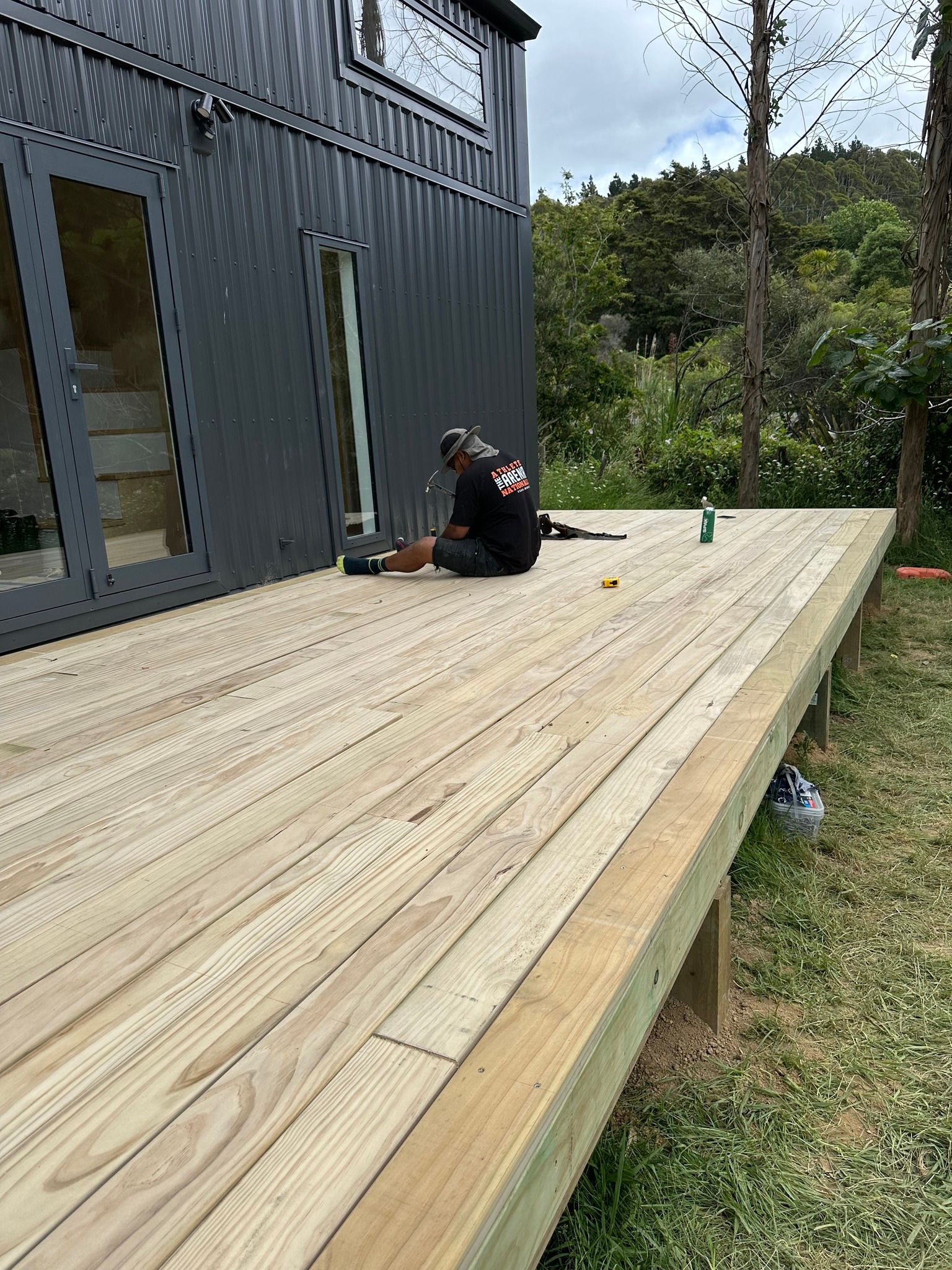 Person working on a wooden deck next to a dark gray house. Person working on a wooden deck next to a dark gray house.