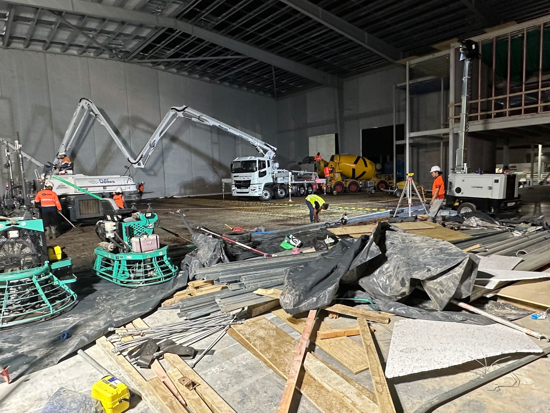 Construction site: concrete pumps, workers, machinery inside a large building, debris on the ground.