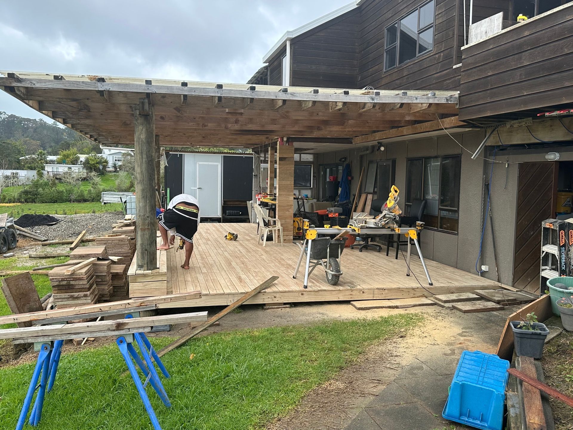 Deck construction in progress; person working on wooden deck with tools. Brown wood and green grass. Deck construction in progress; person working on wooden deck with tools. Brown wood and green grass.