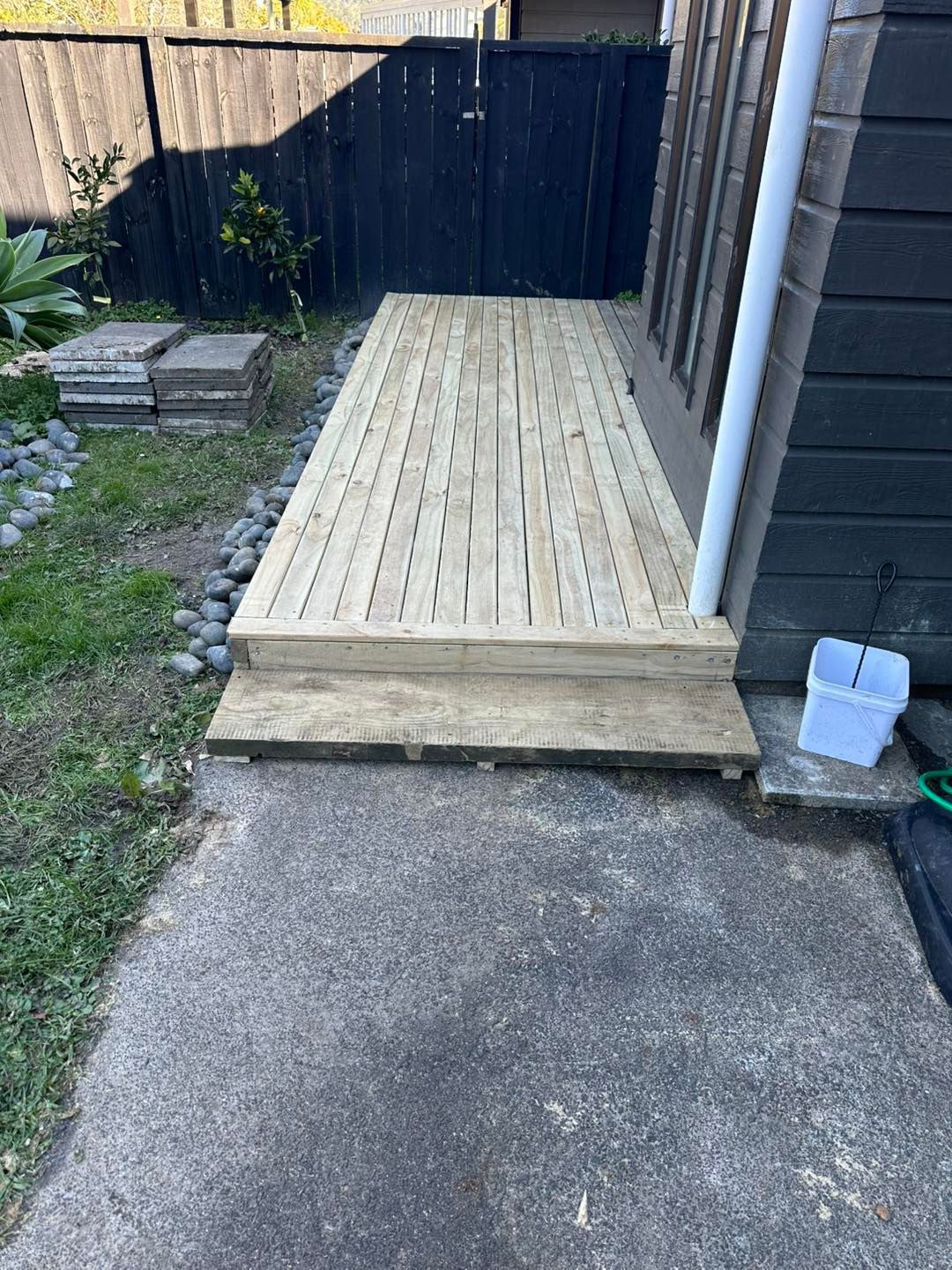 Wooden deck with steps next to a dark wall, accessed from a concrete path.
