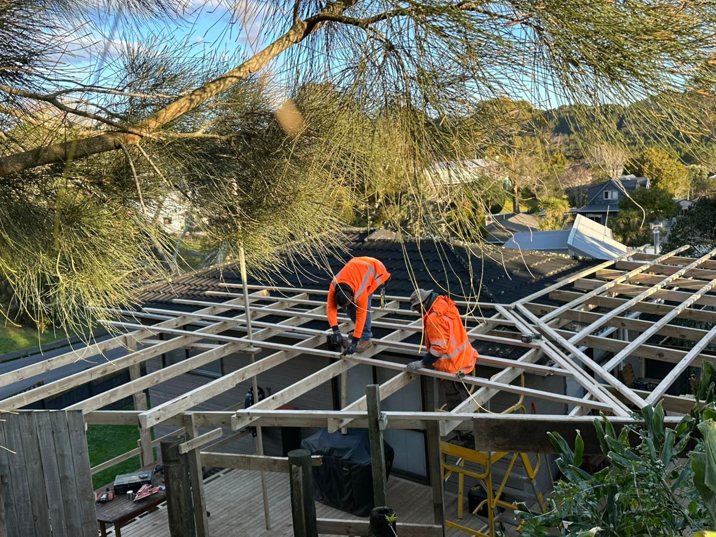 Two workers in orange vests on a roof frame, installing materials.