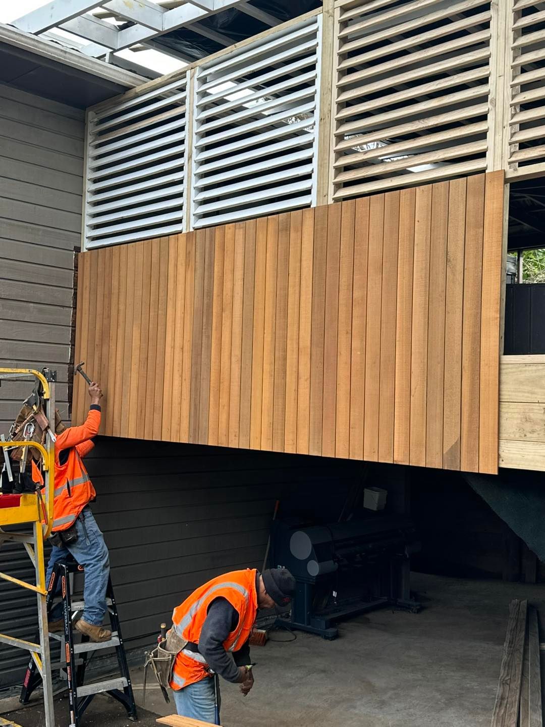 Two construction workers install wooden siding on a building, while the structure has a slat-style window.