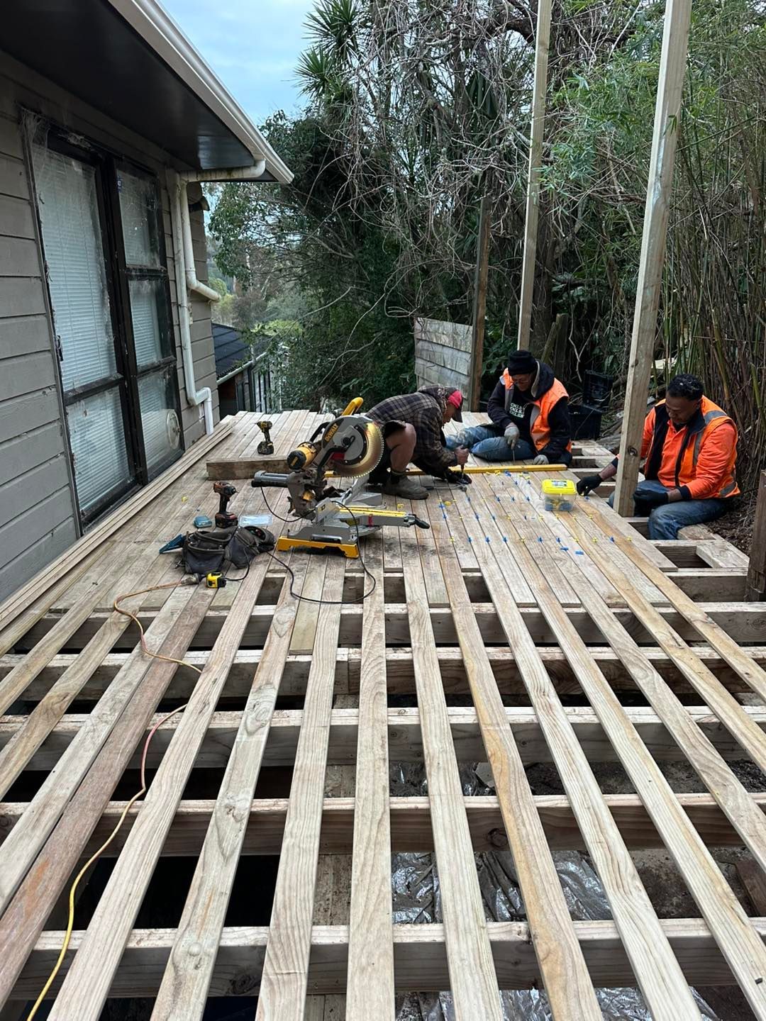 Construction workers building a wooden deck. One uses a saw, others seated nearby.