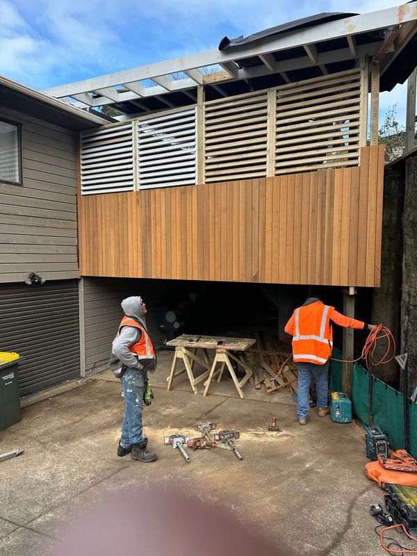Two workers in orange vests at a construction site, under a wooden deck with slatted panels.
