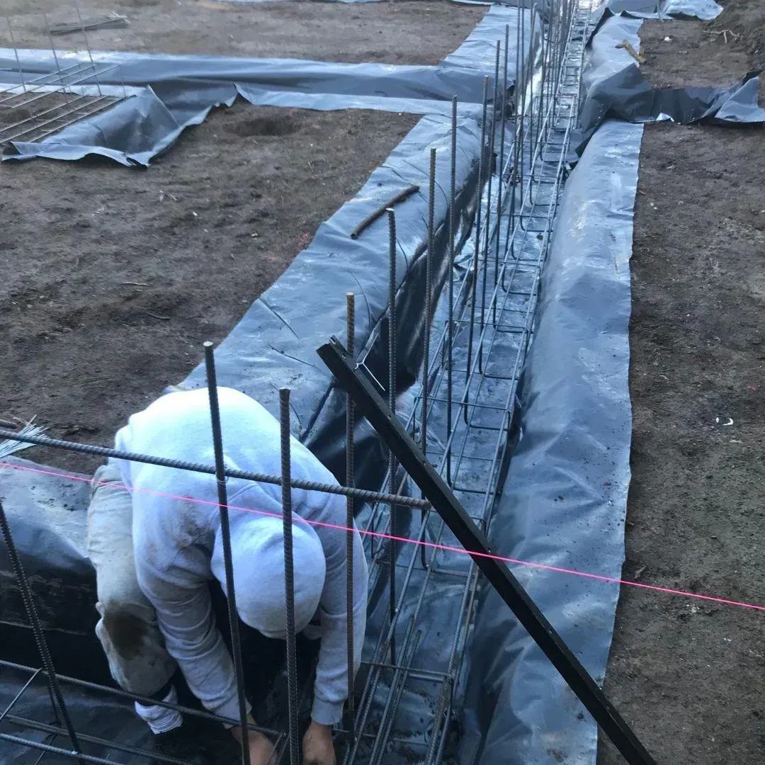 Construction worker installing rebar in a trench. Black plastic lining, gray rebar, brown soil, outdoor setting.