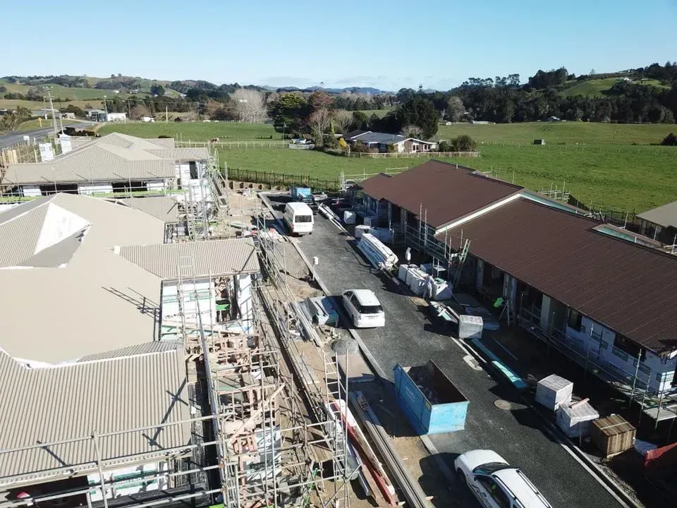 Aerial view of construction site with buildings, vehicles, and green fields in the background.
