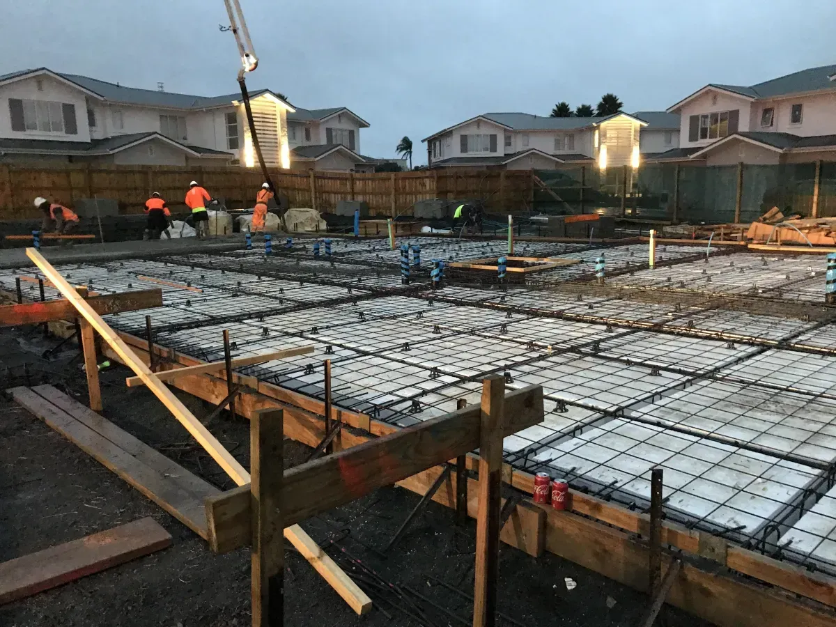 Construction site with workers pouring concrete foundation; houses in background.
