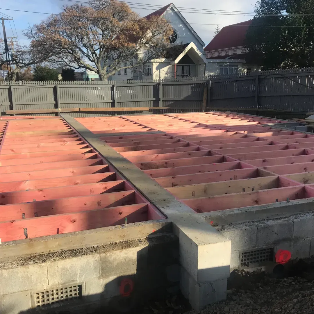 Wooden frame foundation with pink insulation, surrounded by a concrete block base, with a fence and houses in the background.