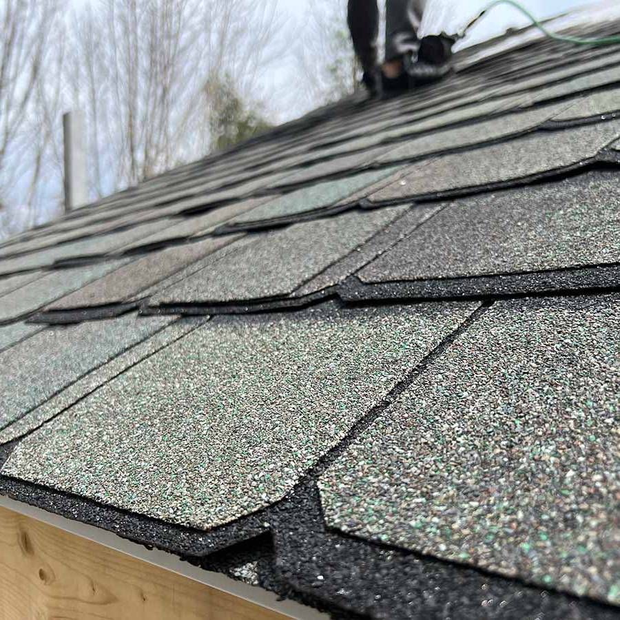 Close-up of a shingled roof with a person working; light and dark grey shingles, overcast sky.