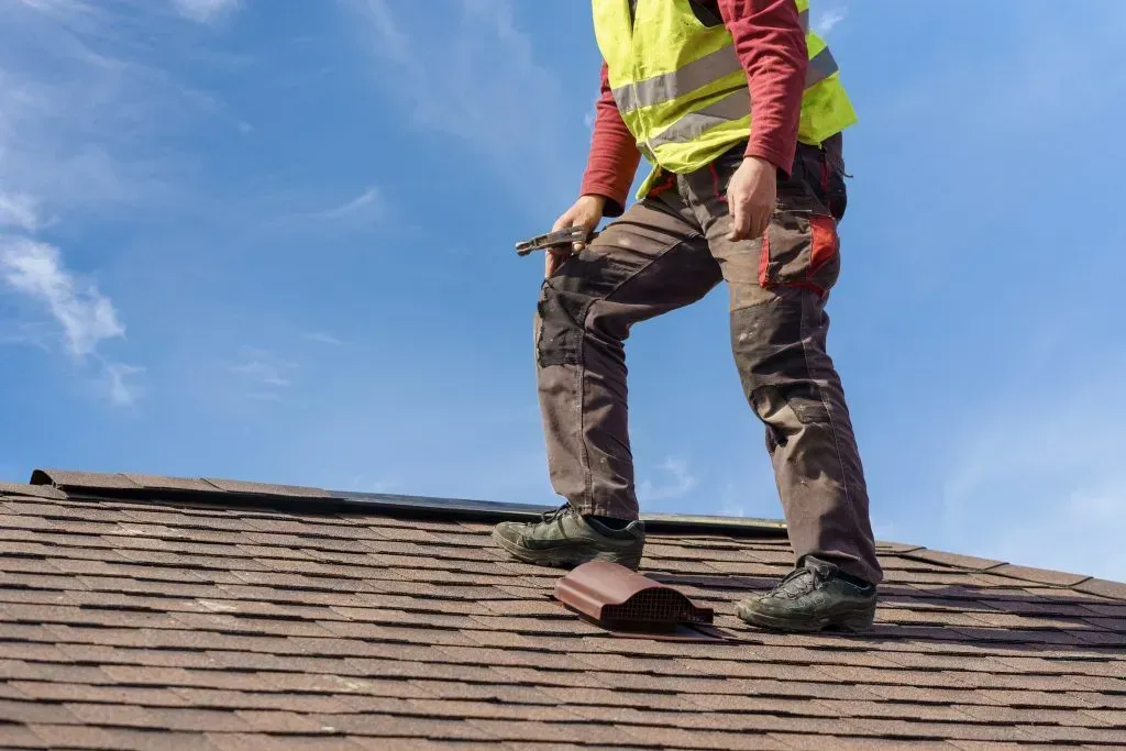 Roofer in safety vest on a brown shingle roof, holding tool, blue sky background.