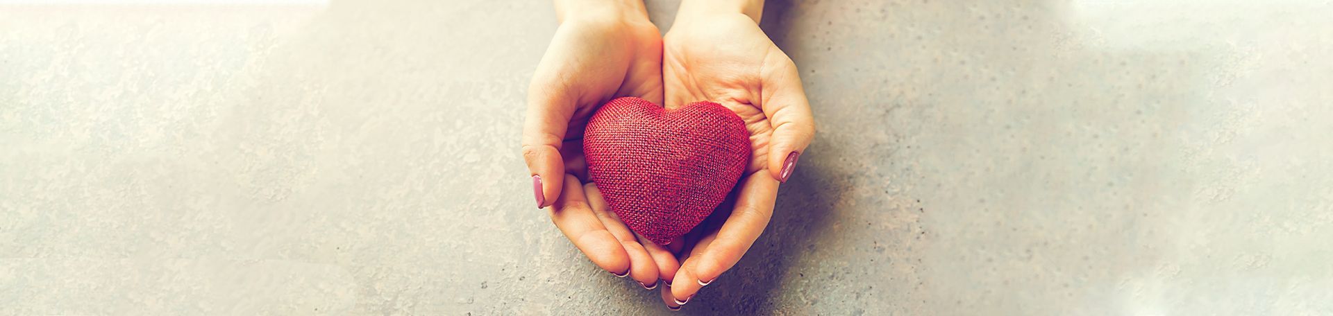 Hands cradling a textured, red heart on a light gray surface.