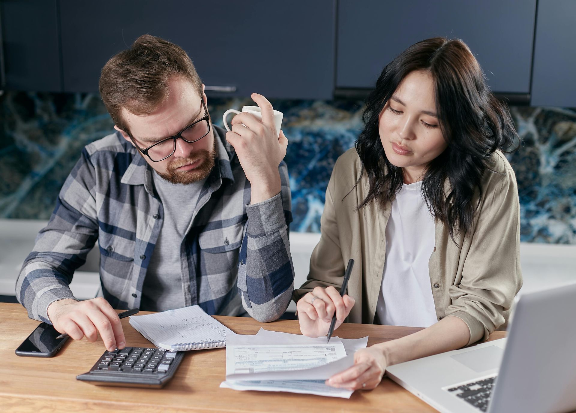 Two coworkers reviewing paperwork at a desk, using a calculator and laptop in an office.
