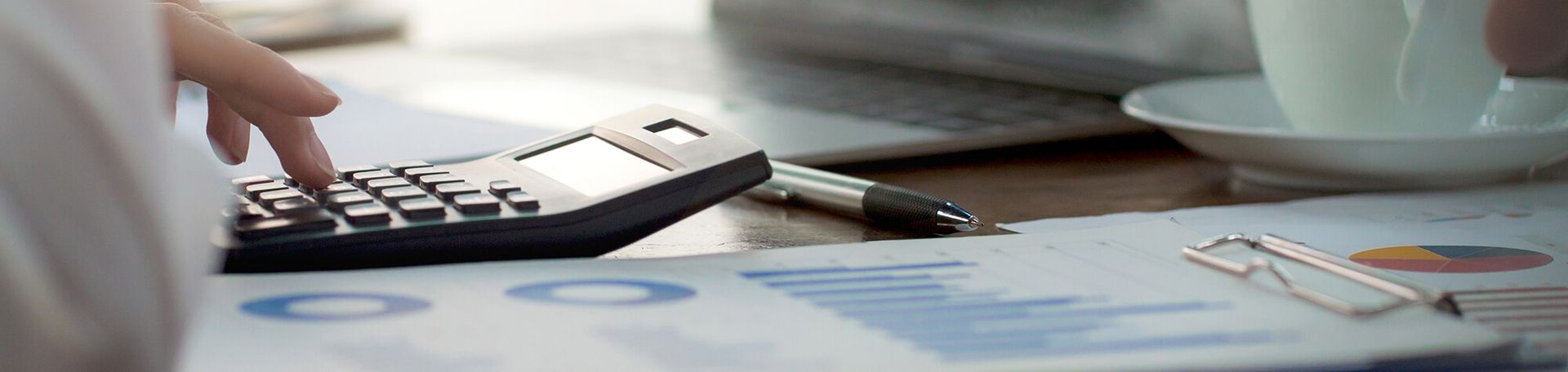 Person's hands using a calculator on a desk with a pen, papers, and a cup of coffee.