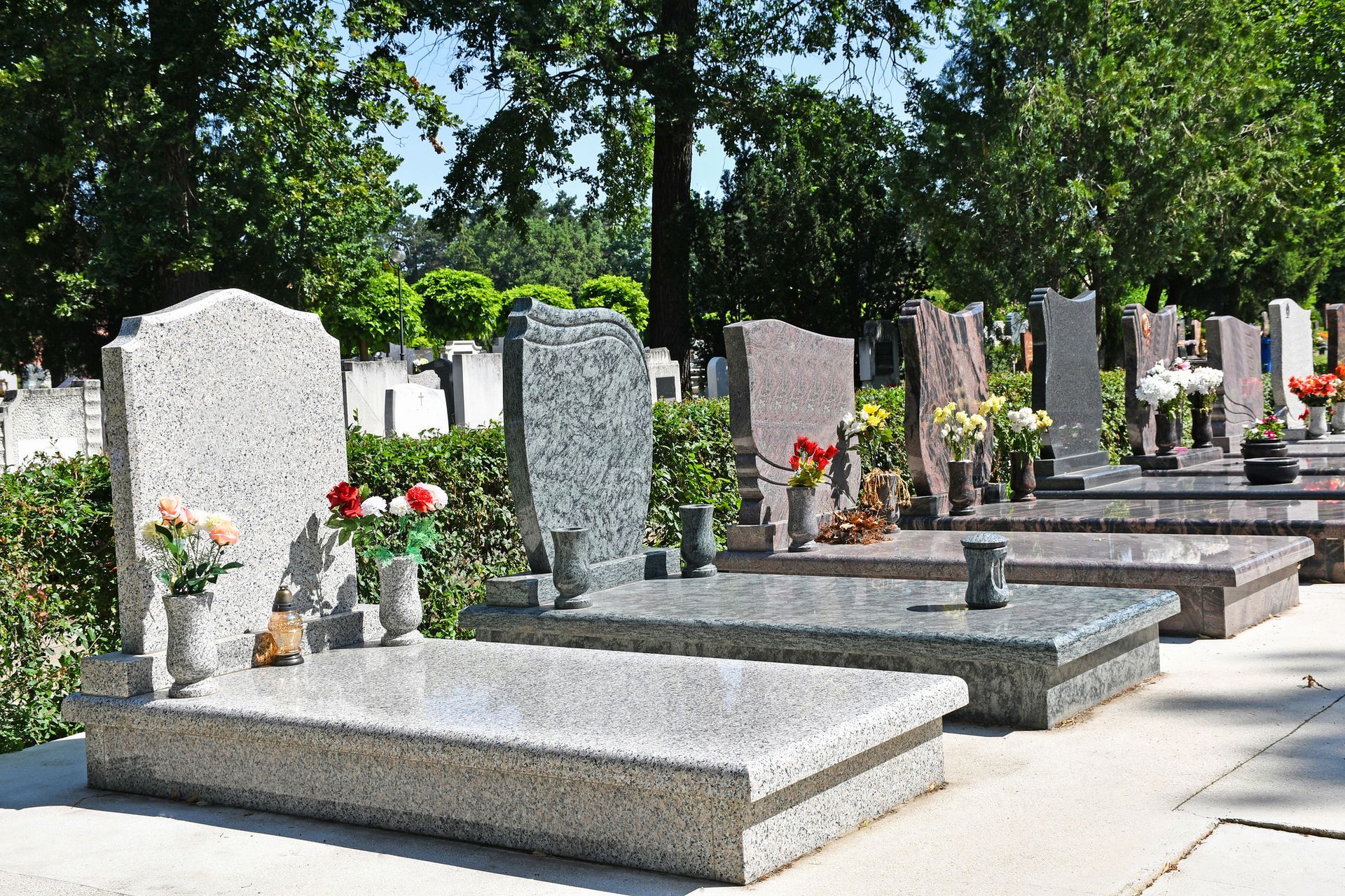 Granite headstones with flower vases in a peaceful cemetery lined with trees on a sunny day scene.