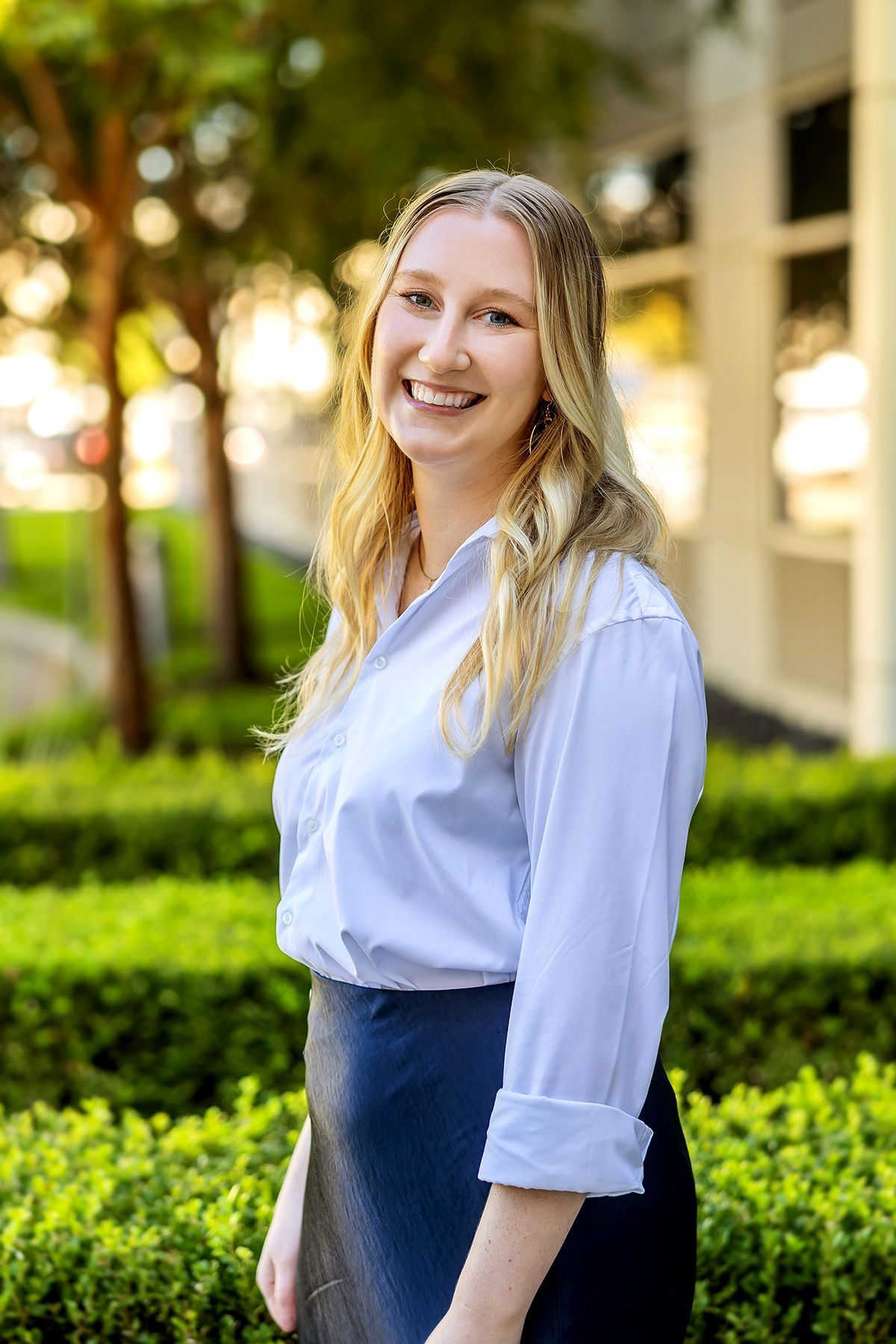 Woman with blonde hair smiles in a white shirt and navy skirt, standing outside near green bushes.