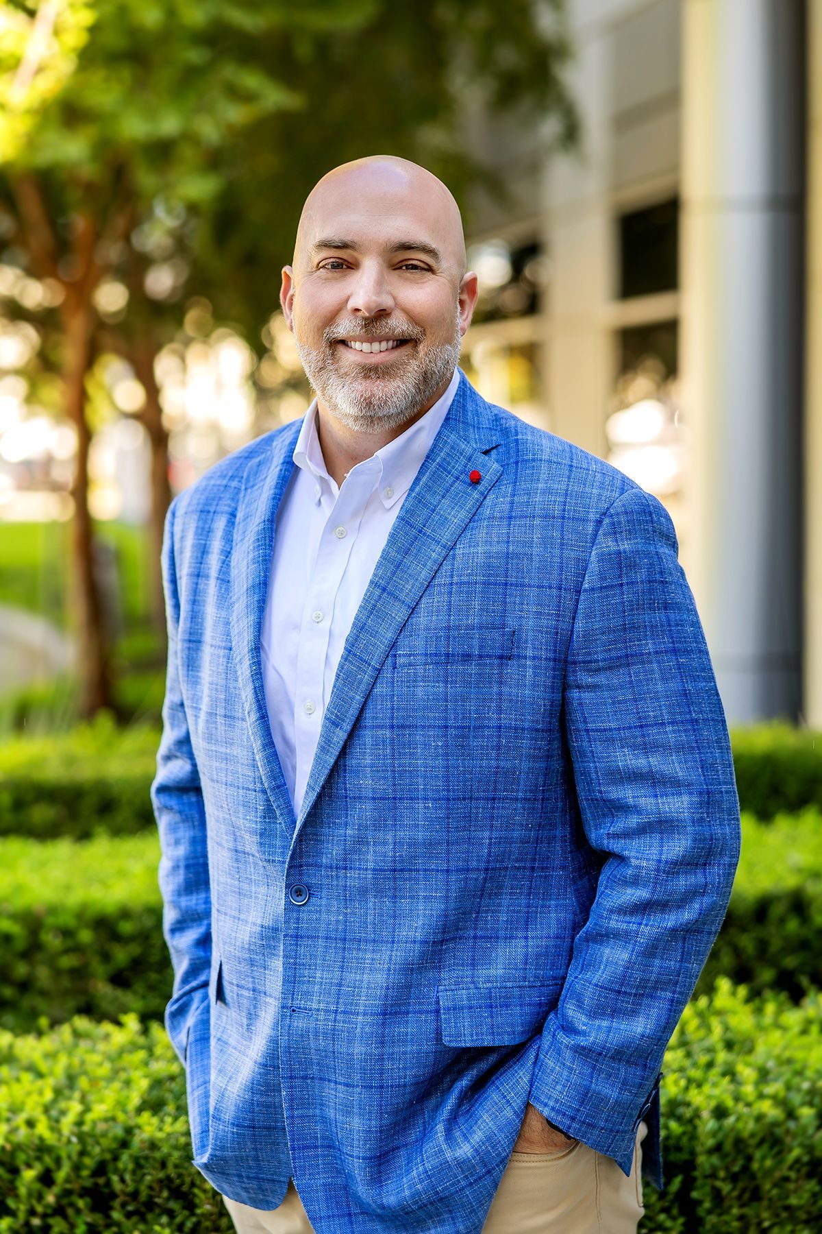 Bald man in blue blazer smiles, hands in pockets, outside a building.
