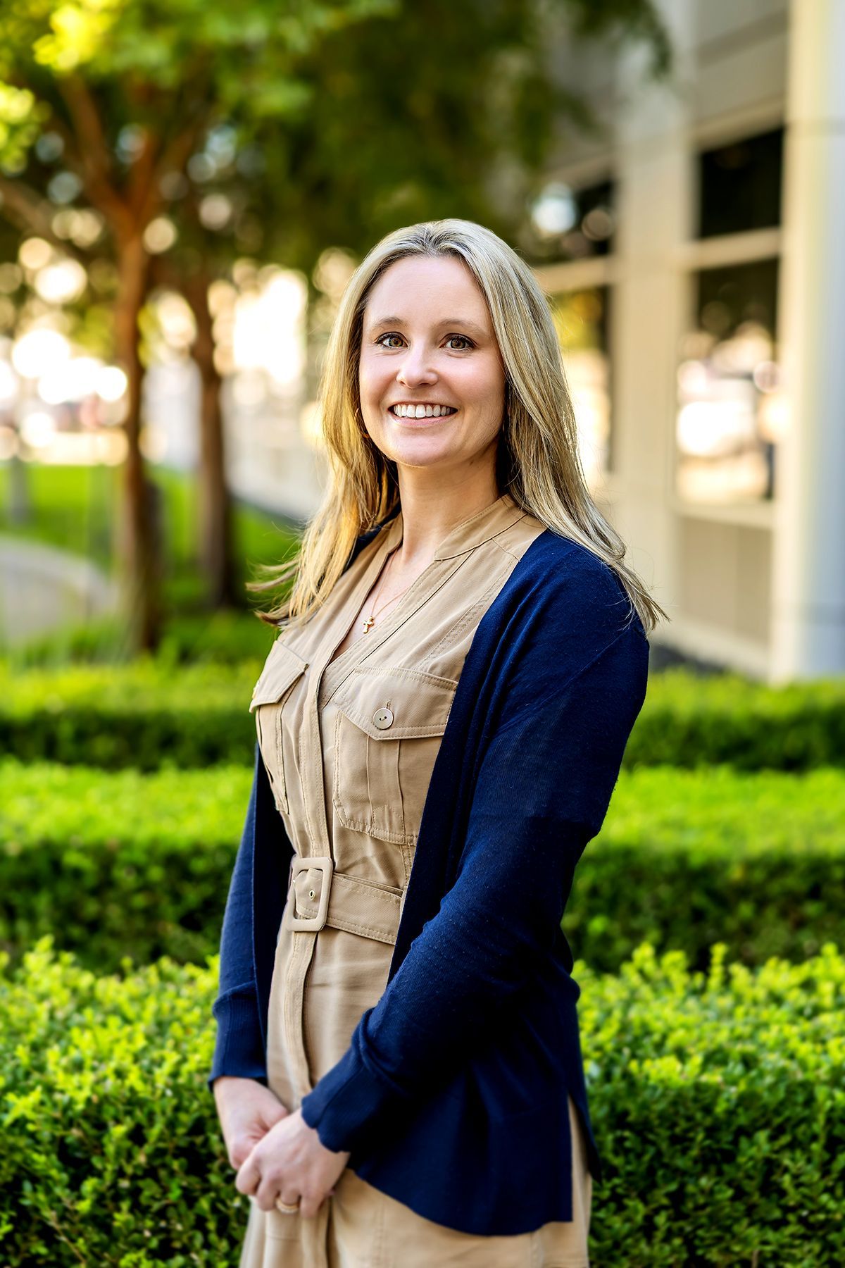 Woman with blonde hair, wearing a beige dress and blue cardigan, smiling outdoors.