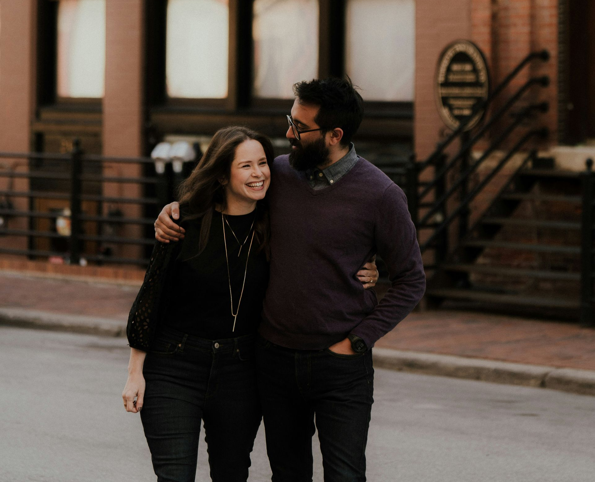 Man with beard and woman smiling, arm in arm, walking on a street, buildings in background.