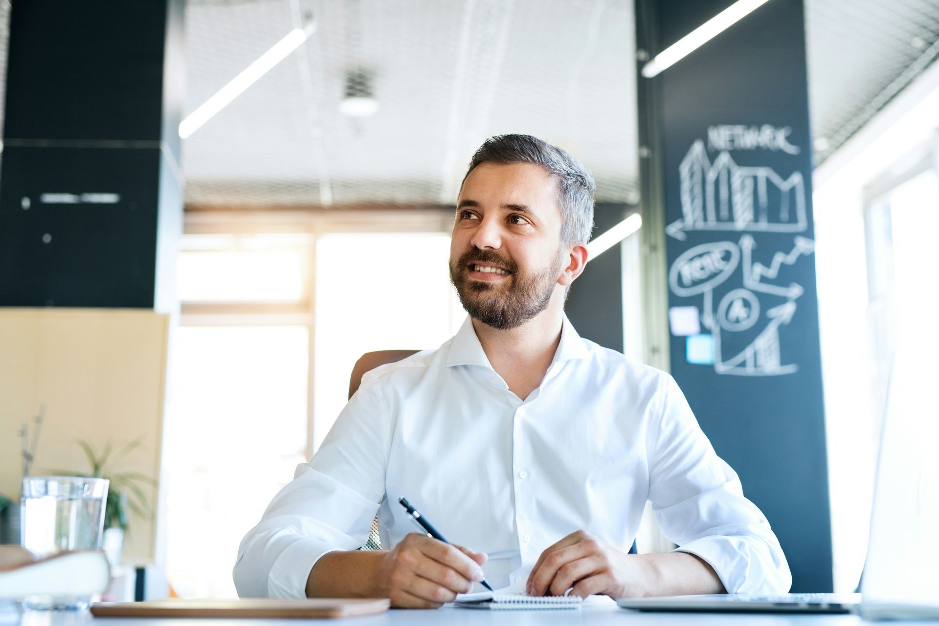 Man with a beard in a white shirt, smiles while holding a pen at a desk in an office.