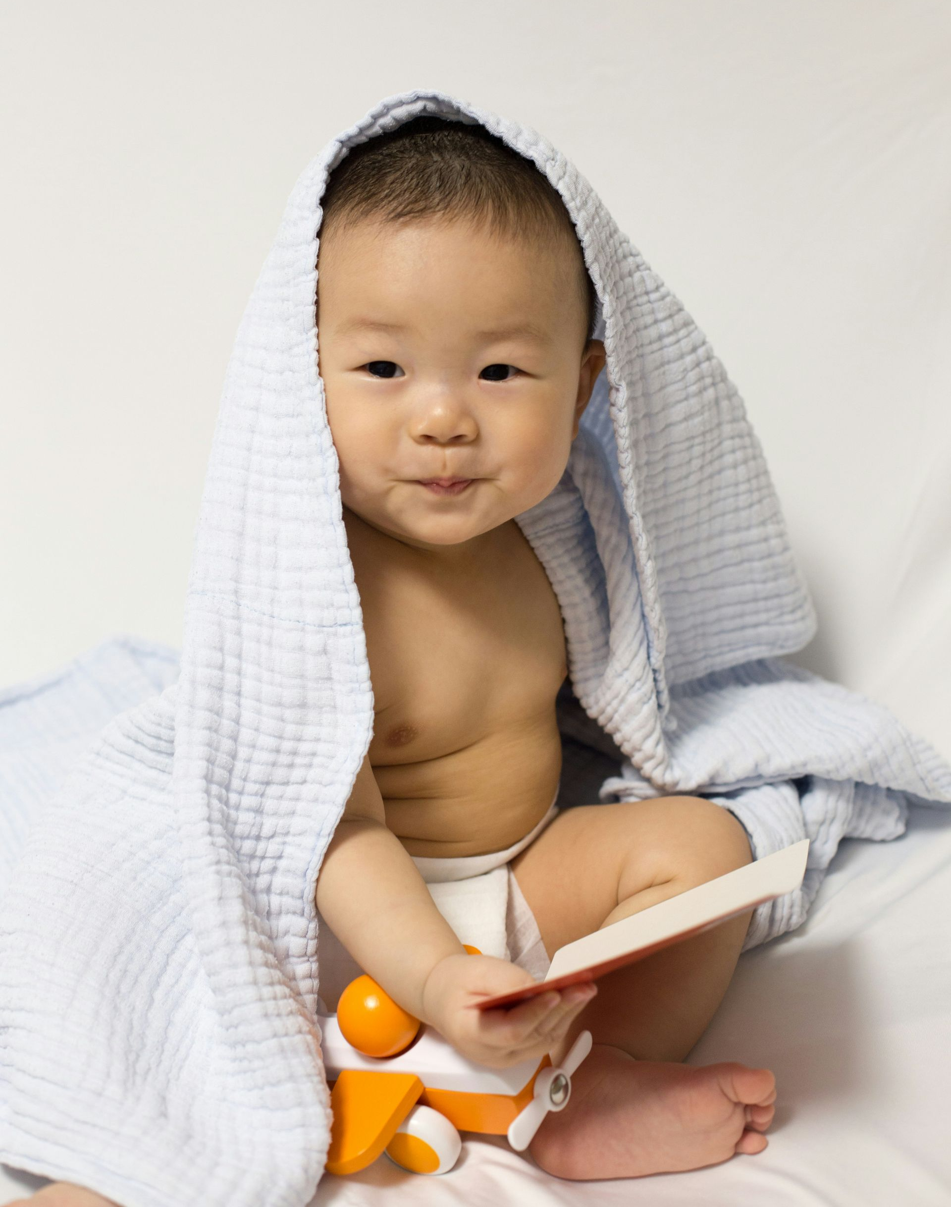 Baby wrapped in a blue towel, holding a book and toy airplane, smiling, seated.