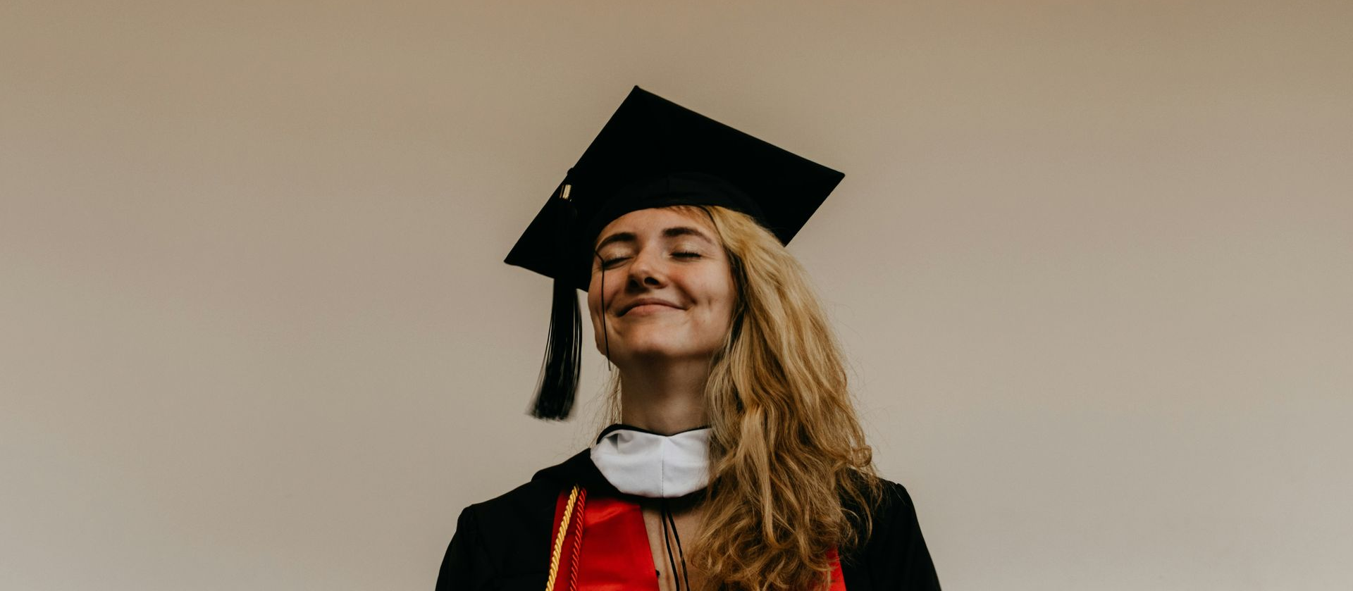 Person in graduation cap and gown smiles with eyes closed against a neutral background.