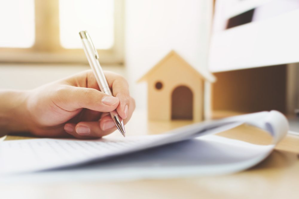 A hand uses a silver pen to sign a document, with a small wooden house model in the background — Murphy Finance Solutions–Darwin & Palmerston In Zuccoli, NT