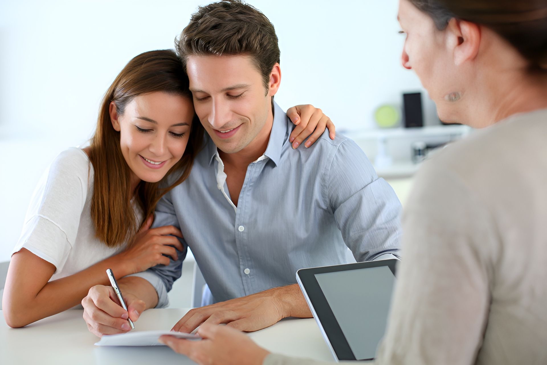 A couple smiles while sitting at a table with a professional, with one person signing a document near a digital tablet — Murphy Finance Solutions–Darwin & Palmerston In Zuccoli, NT