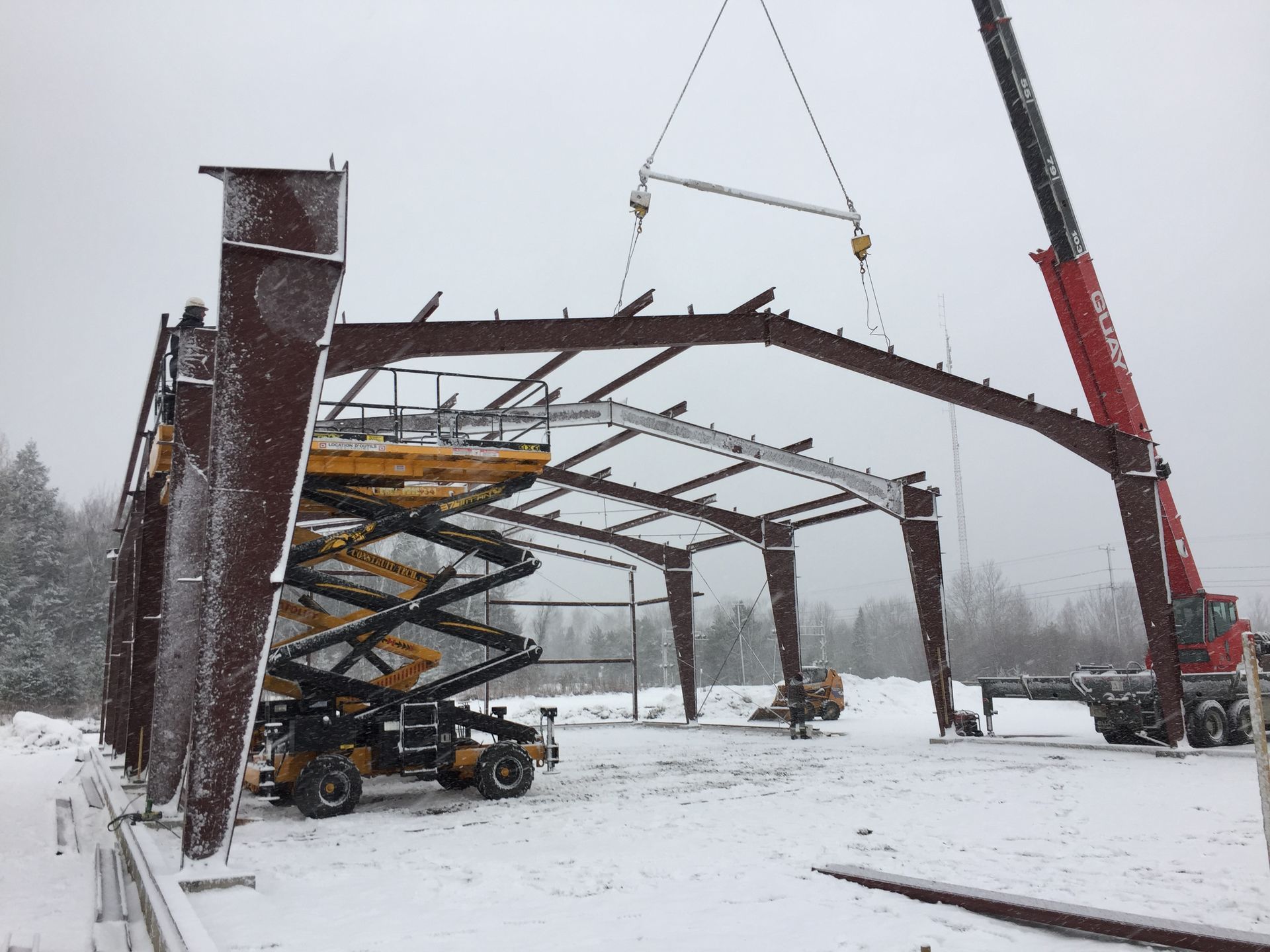 Chantier de construction avec grue soulevant une poutre d'acier pour une charpente de bâtiment dans des conditions enneigées.